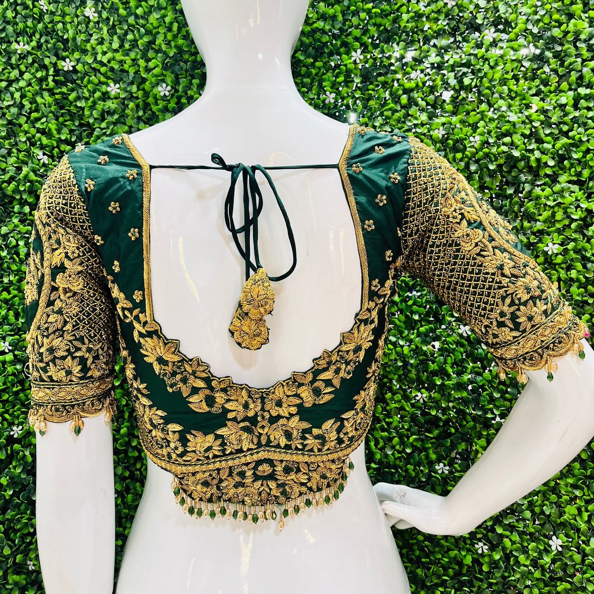 A white mannequin torso wearing a dark green and gold heavily embroidered blouse, photographed from the back against a green foliage wall.