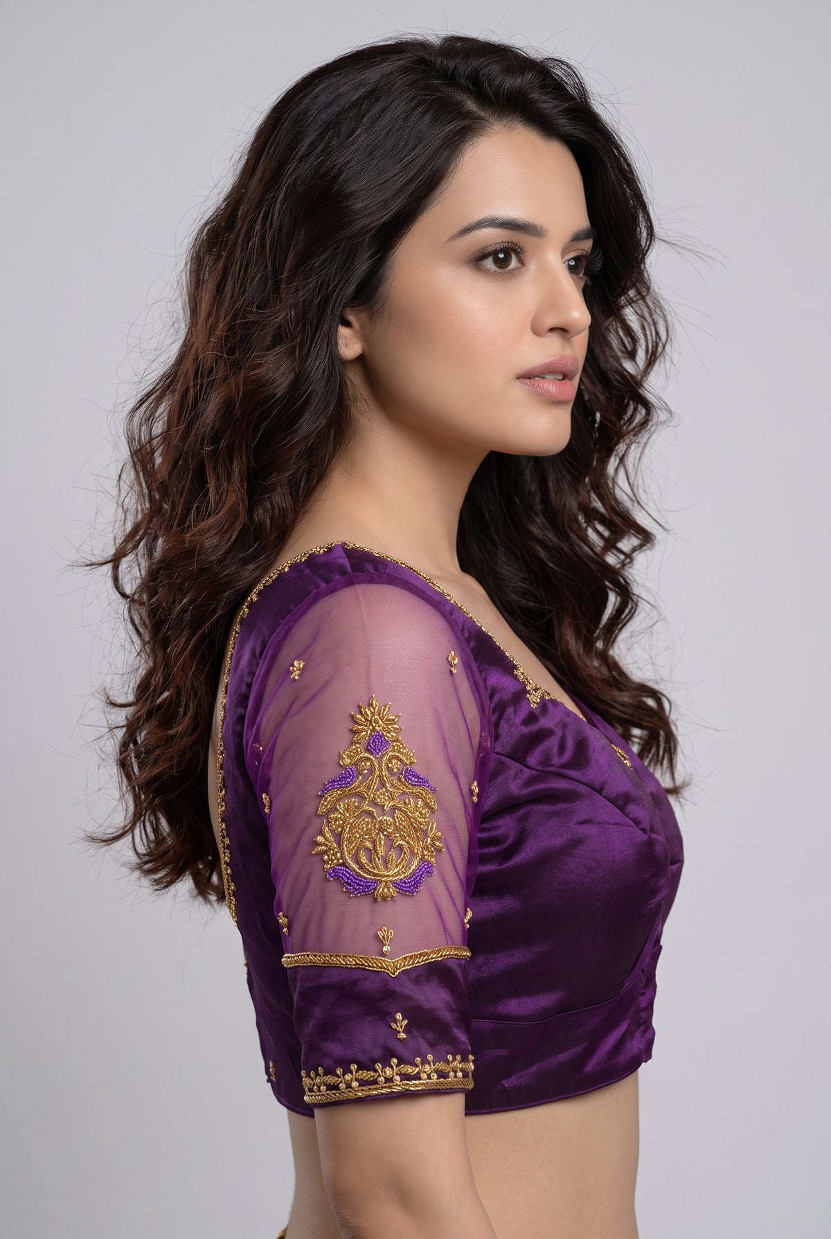 A woman shown in side profile wearing a purple embroidered blouse with sheer mesh sleeves and gold detailing, against a plain light gray studio background.