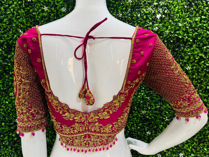 A close-up back view of a bright pink and gold embroidered blouse displayed on a white mannequin torso, with a green hedge background.