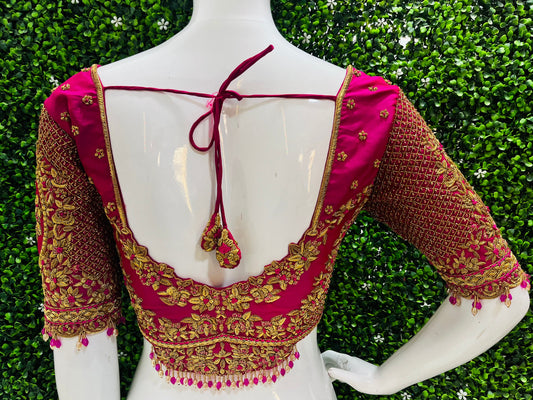 A close-up back view of a bright pink and gold embroidered blouse displayed on a white mannequin torso, with a green hedge background.