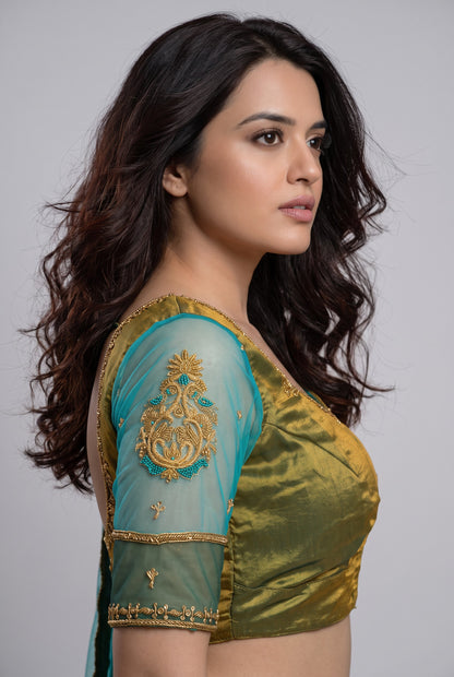 A woman shown in side profile wearing a teal and olive green embroidered blouse with gold detailing, against a plain light gray studio background.
