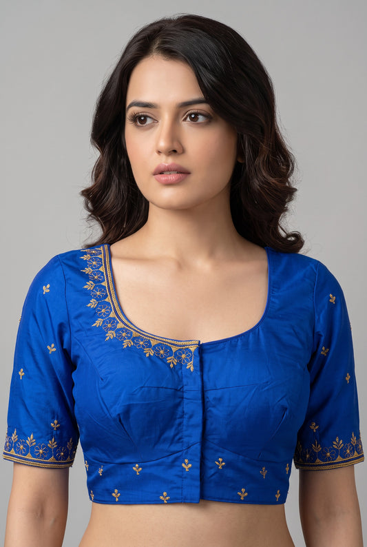 A woman wearing a royal blue short-sleeve blouse with gold floral embroidery around the neckline and sleeve cuffs, photographed against a plain light gray studio background.