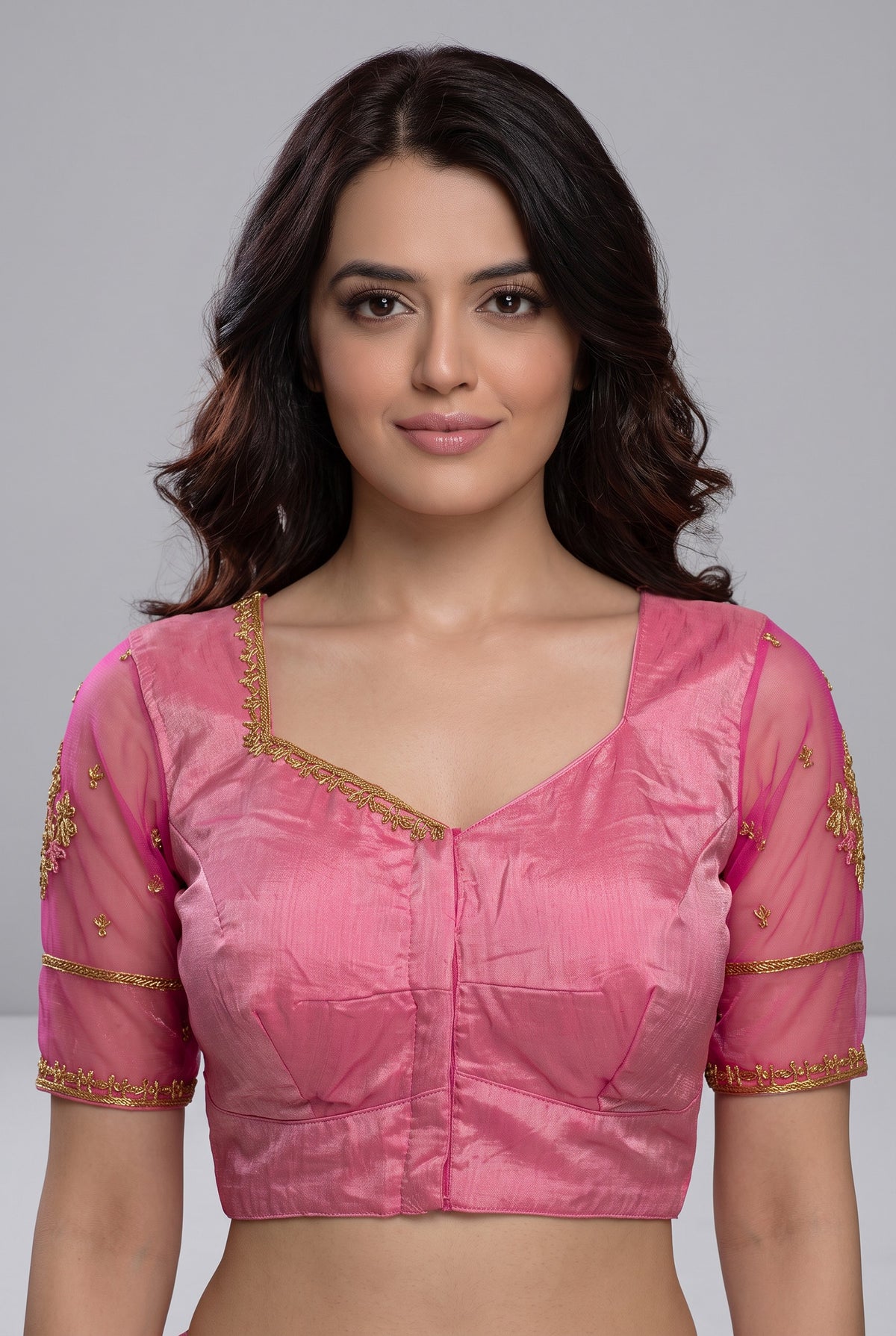 A woman wearing a pink cropped blouse with gold embroidery and sheer sleeves, shown against a plain gray studio background.