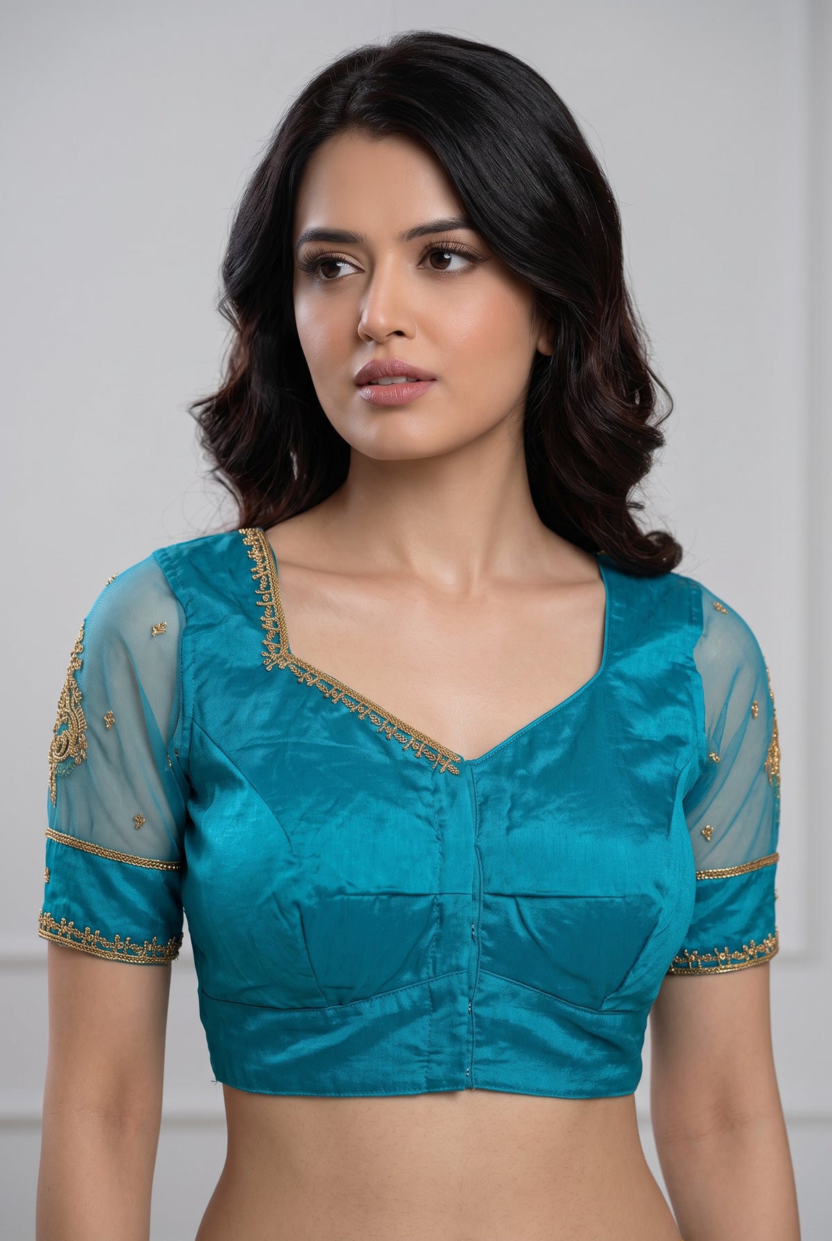 A woman wearing a teal blue satin blouse with gold embroidered details on the neckline and sleeves, paired with sheer mesh sleeves.
