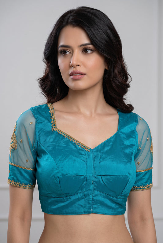 A woman wearing a teal blue satin blouse with gold embroidered details on the neckline and sleeves, paired with sheer mesh sleeves.