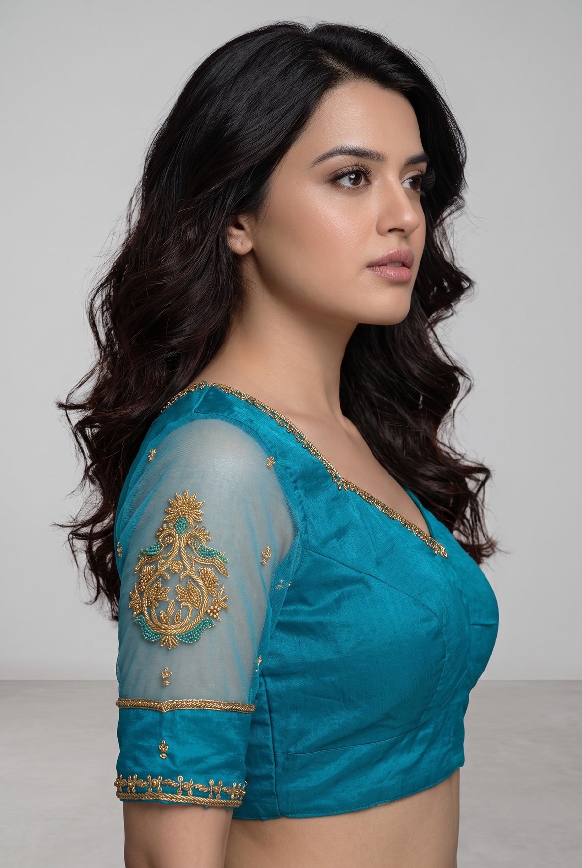 A woman shown in side profile wearing a teal blue blouse with gold embroidery and sheer sleeves, photographed against a plain light gray studio background.