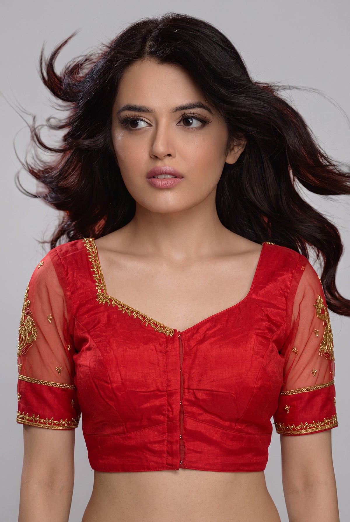 A woman modeling a red sleeveless blouse with sheer mesh sleeves and gold embroidered detailing on the shoulders and cuffs, set against a plain gray studio background.
