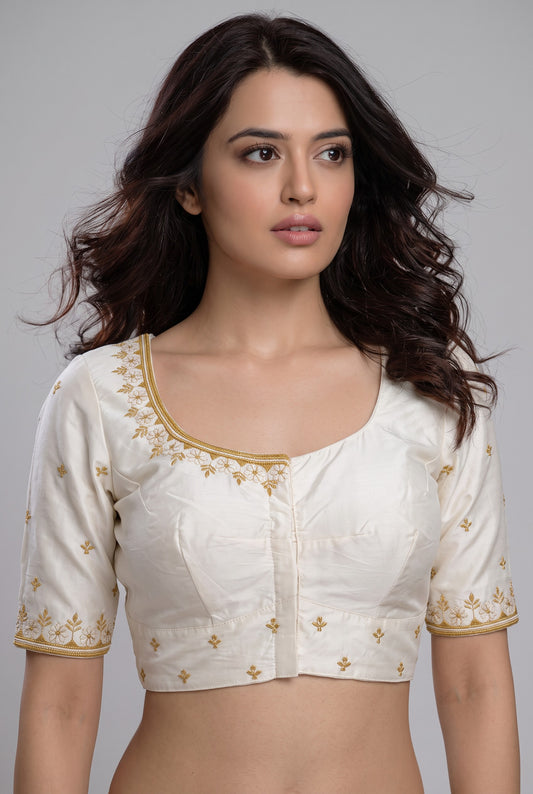 A woman modeling a cream-colored short-sleeve blouse with gold floral embroidery on the neckline, sleeve cuffs, and scattered across the front, set against a solid gray studio background.
