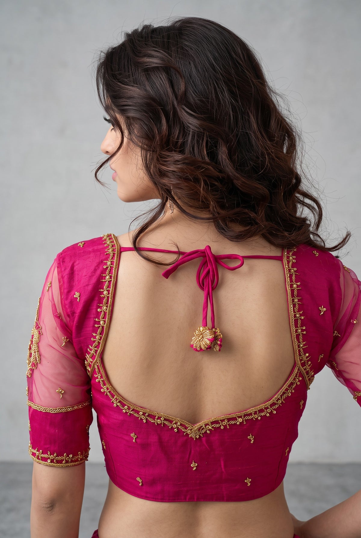 A woman shown from the back wearing a bright pink blouse with gold embroidery and sheer sleeves, tied at the neck with a decorative tassel.
